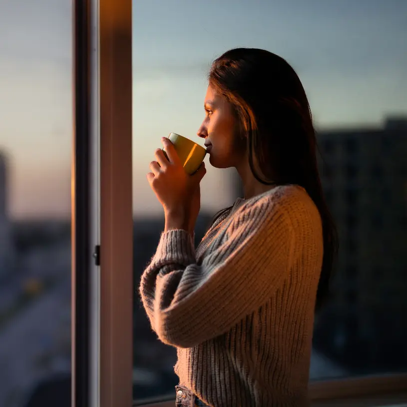 Mujer sentada cerca de una ventana disfrutando una bebida caliente.